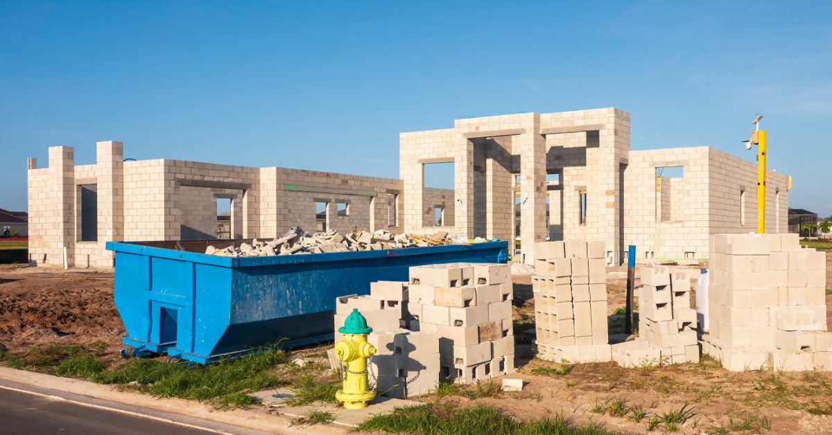 A blue dumpster placed over dirt next to piles of concrete blocks in front of a home structure under construction.